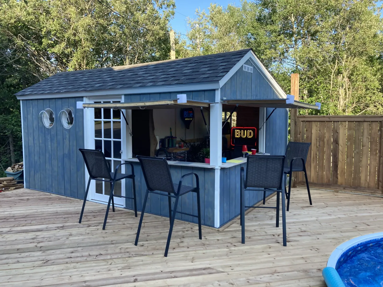 Blue board-and-batten building with a serving hatch propped open on struts over a bar counter, two porthole windows and a glazed door, standing on a timber pool deck in evening light