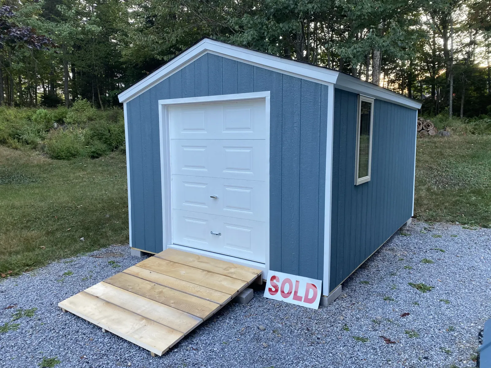 Blue shed with a white overhead garage door and a timber ramp standing on a grass lot with a SOLD sign propped against it