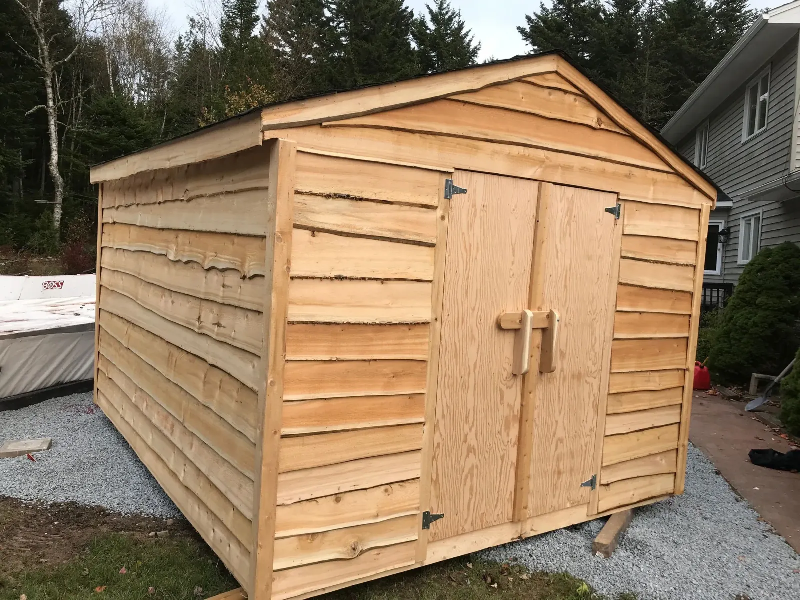 Shed clad in rough-sawn live-edge cedar boards with plywood double doors and a hand-made wooden latch, set on a gravel pad