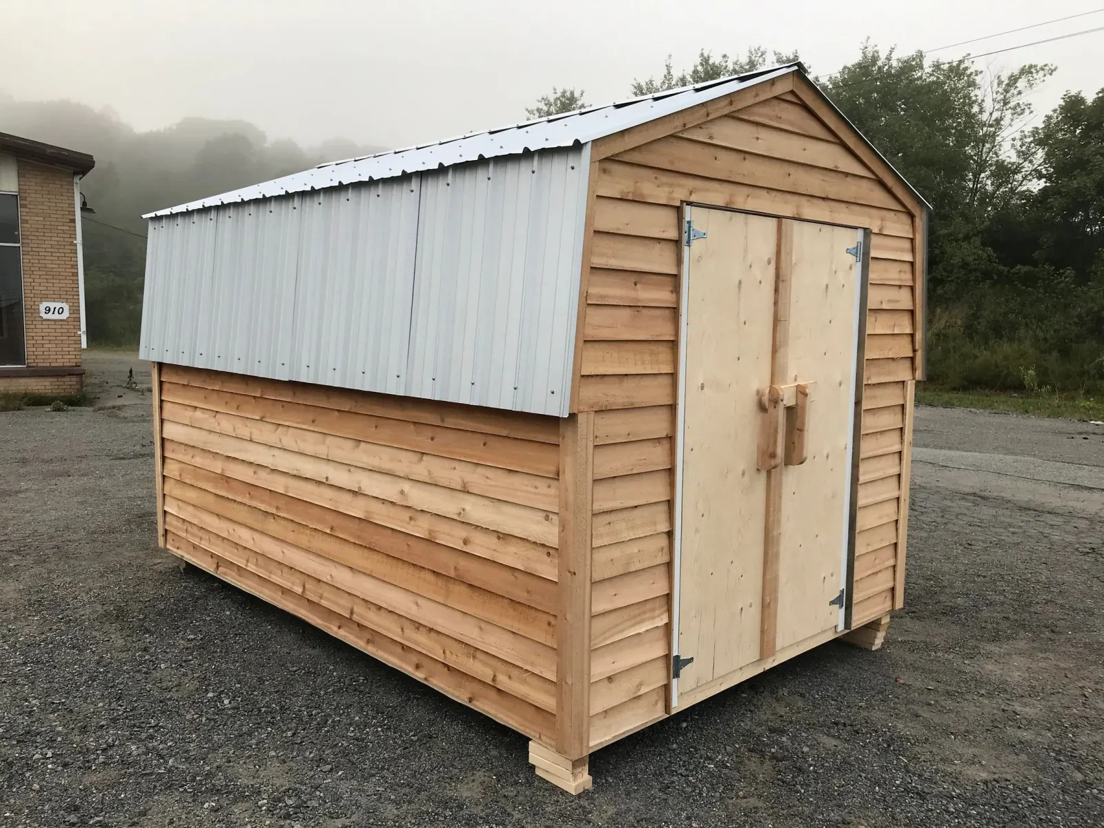 Cedar-sided shed with a galvanized steel roof and plywood double doors, standing on a gravel lot