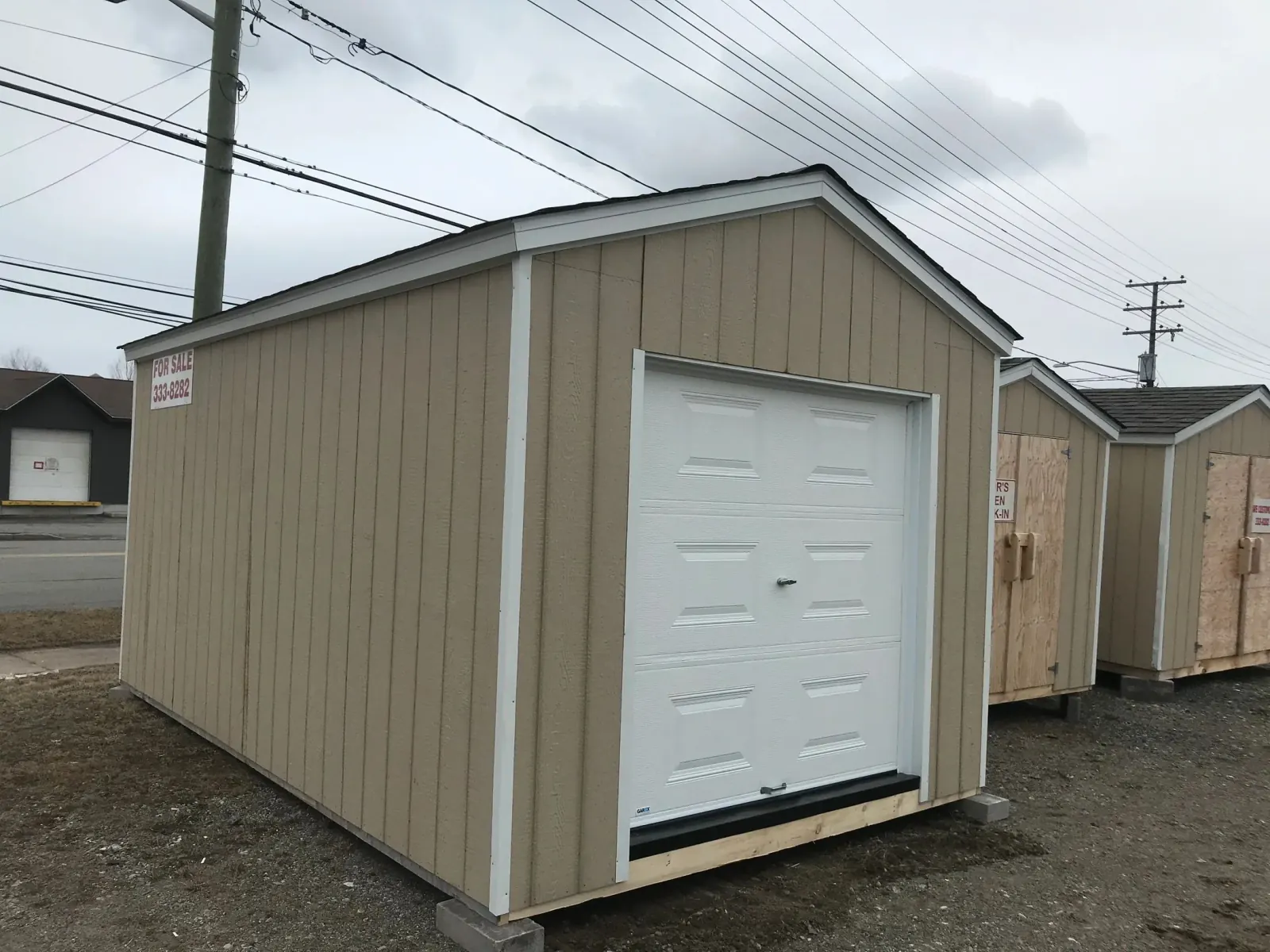 Tan board-and-batten shed fitted with a white overhead garage door, on blocks at the roadside lot