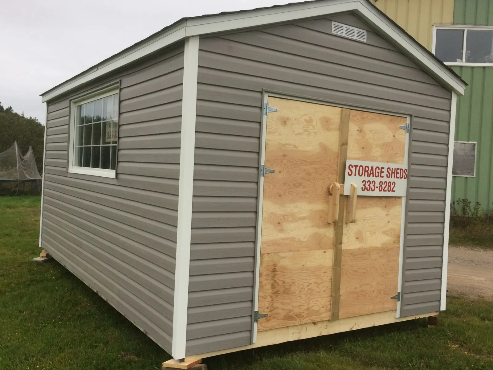 Grey vinyl-sided storage shed with a white-trimmed window and plywood double doors