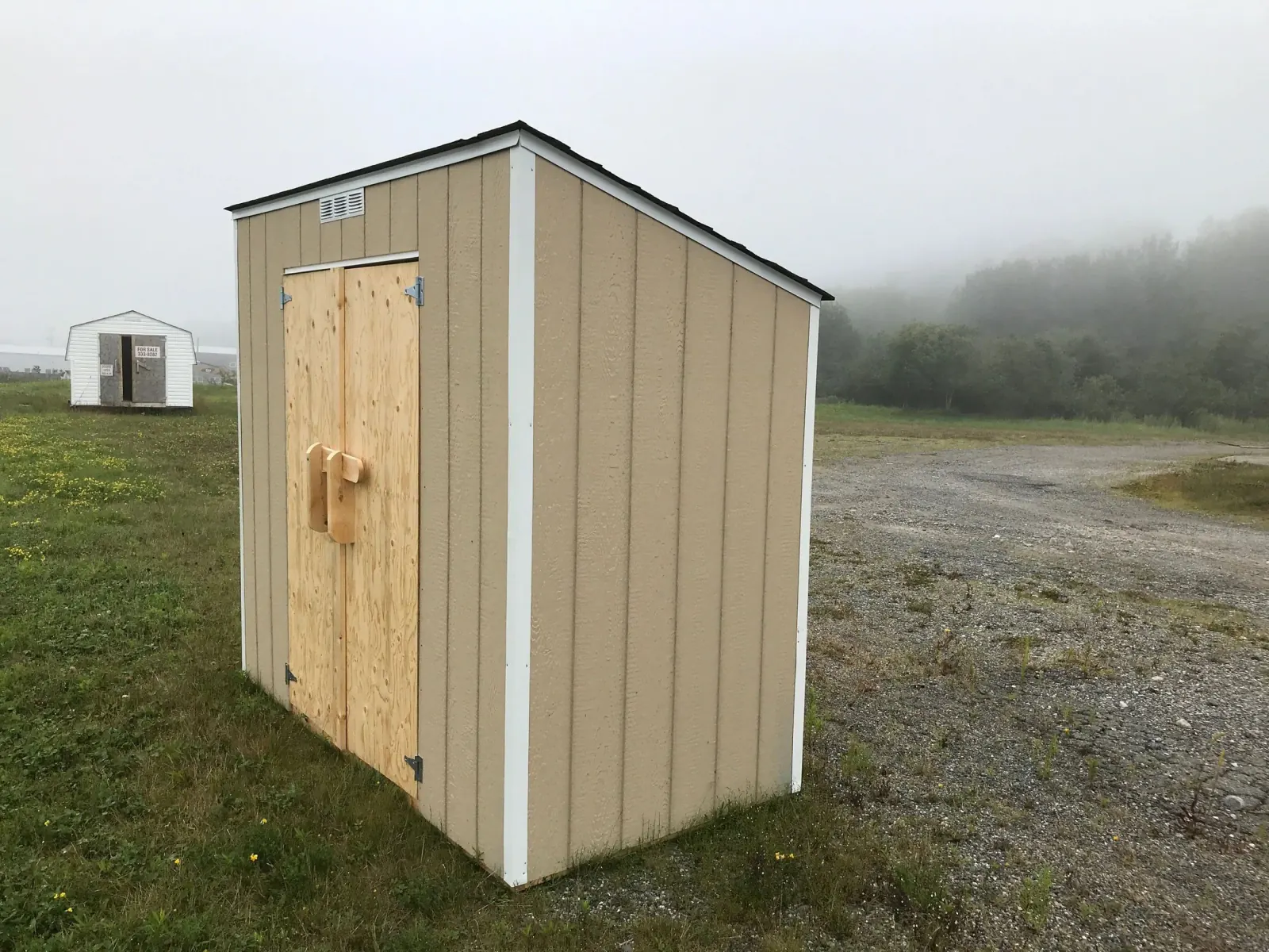Small lean-to roofed shed with plywood doors standing in a grass field on a foggy morning