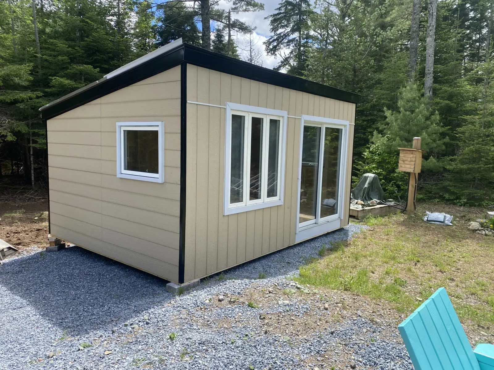 Tan board-and-batten studio building with a black-trimmed mono-pitch roof, a sliding patio door and a three-pane window, set on a fresh crushed-gravel pad at the edge of woods