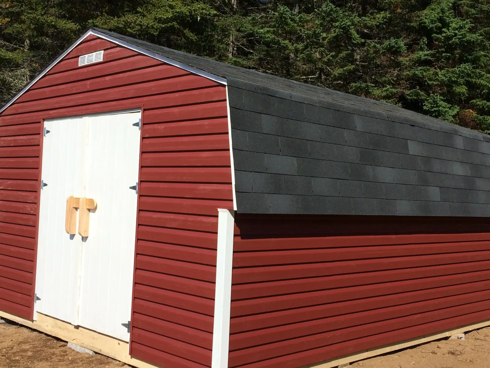 Red barn-style shed with a gambrel roof, white double doors and a wooden turn latch, built against a spruce treeline