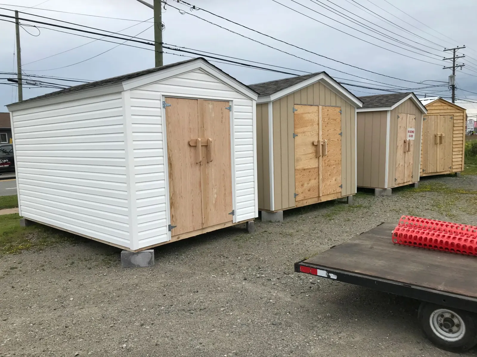 Four finished sheds lined up on a gravel lot, showing white vinyl, board-and-batten and cedar siding side by side