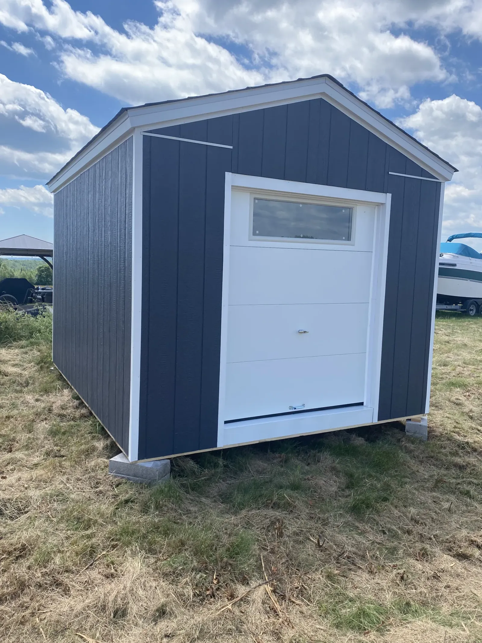 Slate blue-grey shed with white corner trim and a white overhead garage door with a transom window, standing on a grass lot