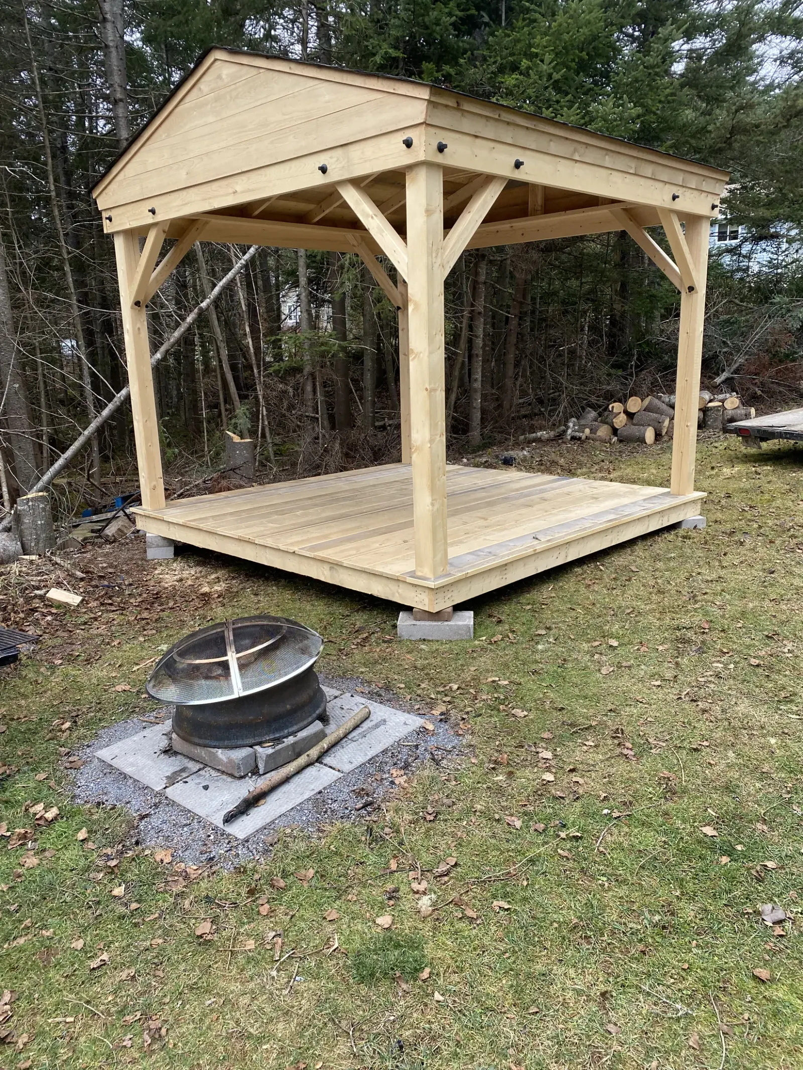 Timber-frame pavilion with heavy posts, braced beams and a new board deck, built on a wooded lot beside a steel fire ring
