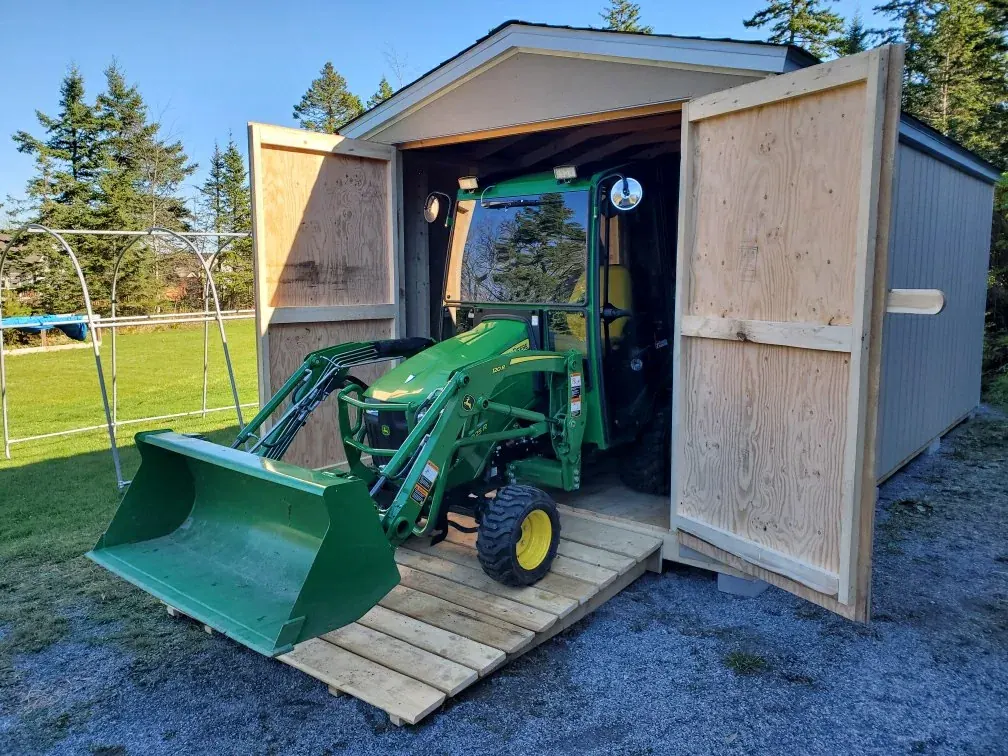 Shed with its plywood double doors open and a green compact tractor with a front loader parked inside on a timber ramp
