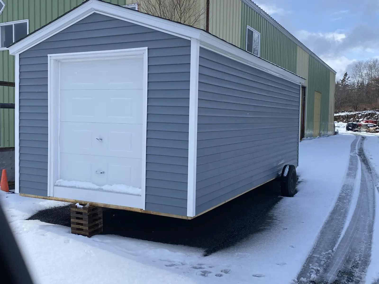 Grey-blue building with a white overhead garage door sitting on a trailer on packed snow beside a workshop
